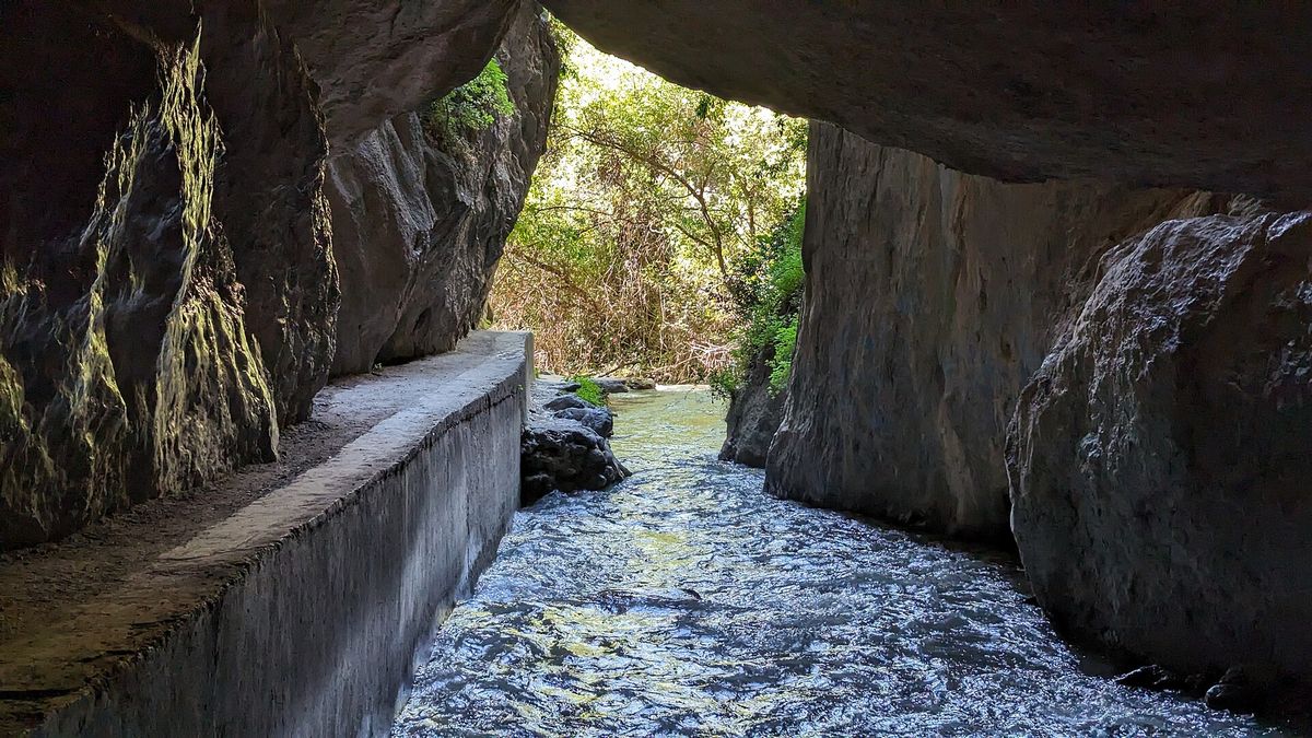Clear water rushes through the narrow rock gorge of Los Cahorros de Monachil beside a concrete path under an overhanging cliff.