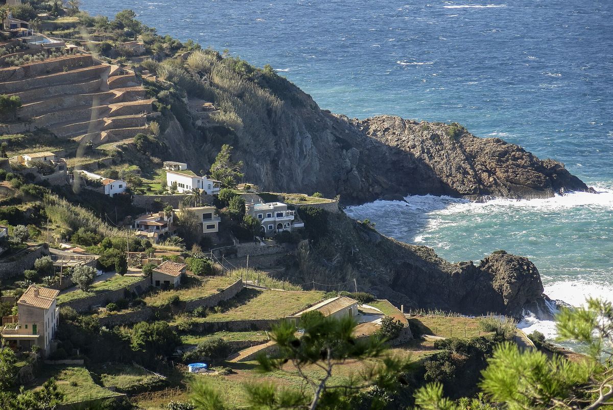Stenterrasserade sluttningar och några få hus leder ner till en klippig udde och det blå havet på kusten av Serra de Tramuntana nära Banyalbufar.