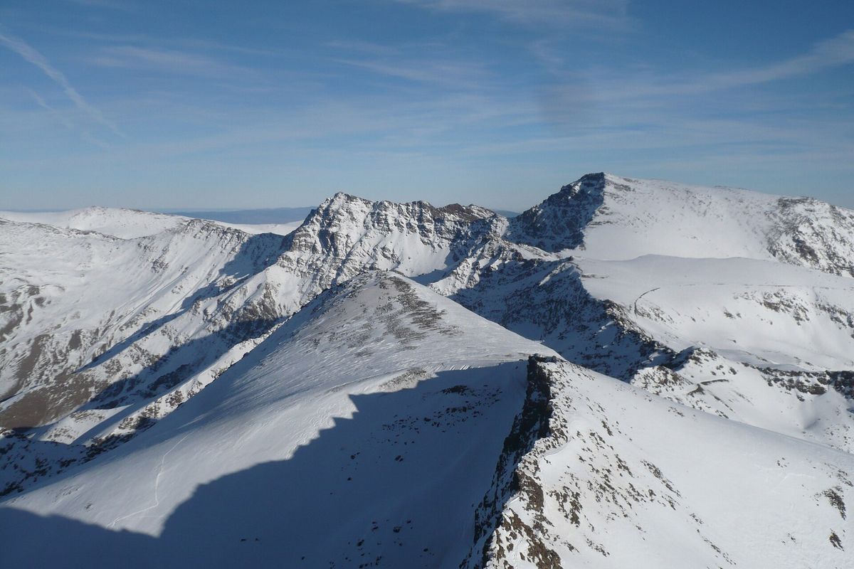Snødekte rygger og topper i Sierra Nevada, blant dem kuppelen på Mulhacén, hever seg under en blek blå himmel.
