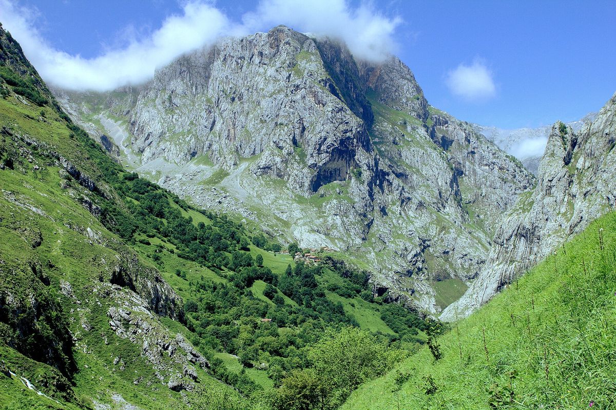 En bratt grønn dal i Picos de Europa med en liten grend nederst under en enorm grå kalksteinsvegg, under blå himmel med lave skyer.