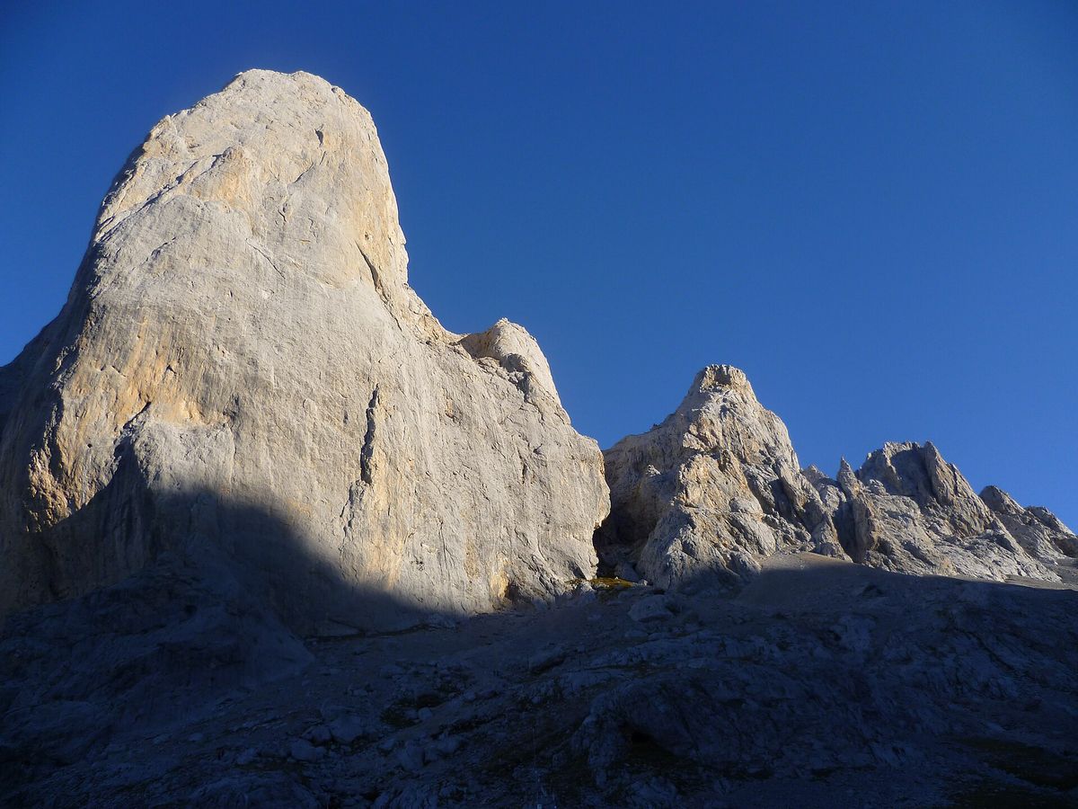 Det bleka kalkstenstornet Picu Urriellu, även kallat Naranjo de Bulnes, reser sig mot en djupblå himmel över stenig mark nära stugan.