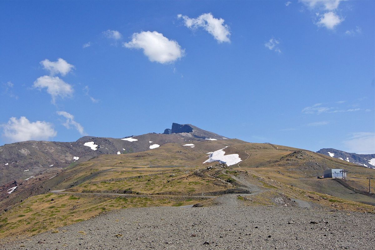 The dark rocky summit of Pico Veleta rises behind brown slopes with patches of snow and gravel tracks under a blue sky with small clouds.